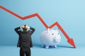 Back view of stressed businessman with falling red chart arrow with piggy bank on blue background. Economic recession, Royalty Free Stock Photo