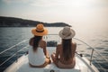 Back view of two young women in swimsuits and hats sitting on the back of the boat and looking at the sea, Friends chilling on a