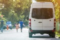 Back view of white passenger medium size commercial luxury minibus van parked n shadow of green tree on summer city street i with Royalty Free Stock Photo