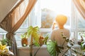 Back view woman drinking tea and looking at the sunrise or sunset while standing at the window in a room with green Royalty Free Stock Photo