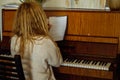 Back view of woman musician with wavy fair hair sitting at old brown piano, holding sheet of score with musical notes.