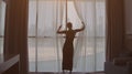 Back view of woman opening curtains and looking out of window to cityscape at hotel in the morning. Modern buildings at Royalty Free Stock Photo