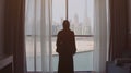 Back view of woman opening curtains and looking out of window to cityscape at hotel in the morning. Modern buildings at Royalty Free Stock Photo