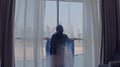 Back view of woman opening curtains and looking out of window to cityscape at hotel in the morning. Modern buildings at Royalty Free Stock Photo