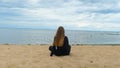 Back view of woman sitting on sandy shore of lake. Concept. Beautiful young woman sitting on sand enjoys view of sea