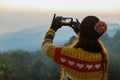 Back view of woman in sweater and thick hat holding smartphone taking photo of mountain view at morning sunrise