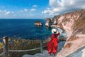 Back view woman wearing a red dress on the edge of a cliff looking at Diamond beach, Nusa Penida. Scenic view of Diamond Beach in