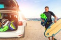 Back view young adult male surfer in wetsuit put in surf board kite equipment on sand beach from van vehicle with Royalty Free Stock Photo