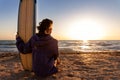 Back view young adult slim sporty female surfer girl with surfboard sitting on sand at ocean coast wave against warm Royalty Free Stock Photo