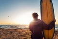 Back view young adult slim sporty female surfer girl with surfboard sitting on sand at ocean coast wave against warm Royalty Free Stock Photo