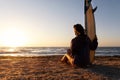 Back view young adult slim sporty female surfer girl with surfboard sitting on sand at ocean coast wave against warm Royalty Free Stock Photo