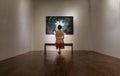 Back view of young asian woman sitting on bench is interested in photograph displayed on white wall at art gallery exhibition hall Royalty Free Stock Photo