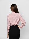 Back view of young brunette woman in formal pink blouse shirt and black tight skirt over grey background