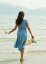 Back view of a young brunette woman in a blue dress walking barefoot on a beach and dangles his feet in the water.
