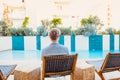 Back view young caucasian man in blue shirt relaxing on chair near swimming pool watching running water and plant flowebed. Summer Royalty Free Stock Photo