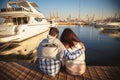 Back view of young couple sitting together on wooden pier in the port with small yachts near to the sea water Royalty Free Stock Photo