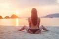 Back view of young female with long brown hair sitting on beach legs spread far apart in black bikini panties admiring