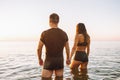 Back view of young fit couple hold their hands with love in sea or ocean at summer sunset. Romantic mood, tenderness