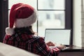 Back view of young freelancer woman in a red santa claus christmas hat sitting near window and working on laptop. Home work Royalty Free Stock Photo