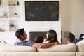 Back view of young Hispanic family of four sitting on the sofa watching TV, mum looking at dad