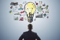 Back view of young male in suit looking at creative business sketch with huge light bulb and other icons. Idea, innovation and Royalty Free Stock Photo