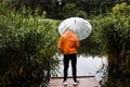 Back view of a young man in an orange raincoat with an umbrella standing on a wooden pier near a lake Royalty Free Stock Photo