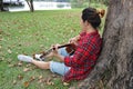 Back view of young relaxed man in red shirt leaning against a tree and playing acoustic guitar in beautiful outdoor park. Royalty Free Stock Photo