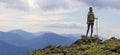 Back view of young slim backpacker tourist girl with stick standing on rocky top against bright blue morning sky enjoying foggy mo