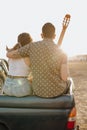 Back view of young travelers lovers having fun with the guitar on the jeep 4x4 car. Couple making a wanderlust vacation on sunset