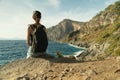 Back view of young woman with backpack sit and rest on rock looking at the sea from rocky coast at sunset Royalty Free Stock Photo