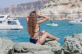 Back view on a young woman in one-piece swimsuit with curly hair sitting on rocks with arms raised looking away Royalty Free Stock Photo