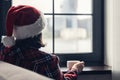 Back view of young woman in a red santa claus christmas hat sitting near window, having breakfast with cup of coffee. Concept. Royalty Free Stock Photo