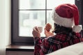 Back view of young woman in a red santa claus christmas hat sitting near window, having breakfast with cup of coffee and croissant Royalty Free Stock Photo