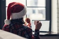 Back view of young woman in a red santa claus christmas hat sitting near window with laptop, having breakfast with cup of coffee Royalty Free Stock Photo