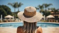 Back view of young woman in straw hat standing near swimming pool at resort