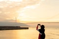 Back view of young woman using smartphone camera for making picture of sunset in ocean at akashi kaikyo bridge, female traveler.