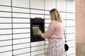 Back view of young woman with wavy fair hair in dress standing at beige self-service delivery locker, touching screen.