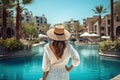 Back view of young woman in white dress and straw hat standing by swimming pool at luxury resort, Happy tourist girl rear view