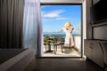 Back view of young woman in white summer dress and straw hat on open balcony terrace of resort hotel room with sea view landscape Royalty Free Stock Photo