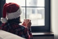 Back view of young woman in a red santa claus christmas hat sitting near window, having breakfast with cup of coffee. Concept. Royalty Free Stock Photo