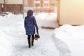 Back of woman in dawn jacket walking through city street during heavy snowfall and blizzard in winter. Bad weather forecast.