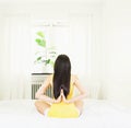 Back of young asian woman wearing yellow undershirt exercising yoga while sitting on a white bed by the window with a thin curtain Royalty Free Stock Photo