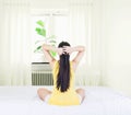 Back of young asian woman wearing yellow undershirt exercising yoga while sitting on a white bed by the window with a thin curtain Royalty Free Stock Photo