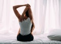 Back of young Caucasian woman stretching as she just woke up from healthy sleep on the bed with the window in background.