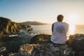 Back of a young traveler sitting on a cliff by the ocean coast watching a beautiful sunset Royalty Free Stock Photo