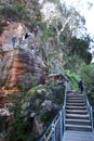 Tourists hiking at steep zigzag trail of Grand Stairway along red rock shear cliff with vegetation in Blue Mountains National Park