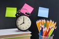 On the background of a black Board with stickers, there is an alarm clock on the books and a glass with colored pencils