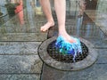 The child`s feet jump on the fountain, illuminated by blue light. A jet of water rises from the surface of the iron lattice. Spray