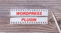 Background for text. White strips of paper with the text WORDPRESS PLUGIN and a white pencil next to it on a wooden background