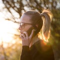 Backlit rear view of young woman talking on cell phone outdoors in park at sunset. Girl holding mobile phone, using Royalty Free Stock Photo